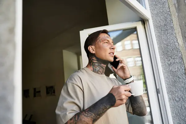 A handsome young man stands by the window, sipping coffee and talking on the phone, relaxed at home. — Stock Photo