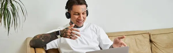 A handsome young man relaxes at home, smiling and chatting on his laptop while wearing headphones. — Stock Photo