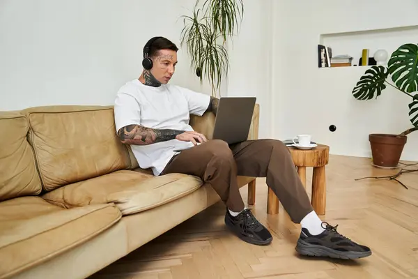 A young man sits comfortably on a couch, focused on his laptop while enjoying music and coffee. — Stock Photo
