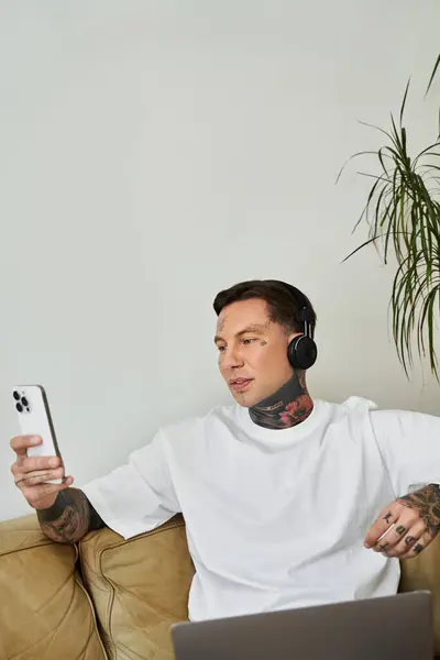 A stylish young man sits comfortably on a couch, enjoying music and scrolling through his phone. — Stock Photo