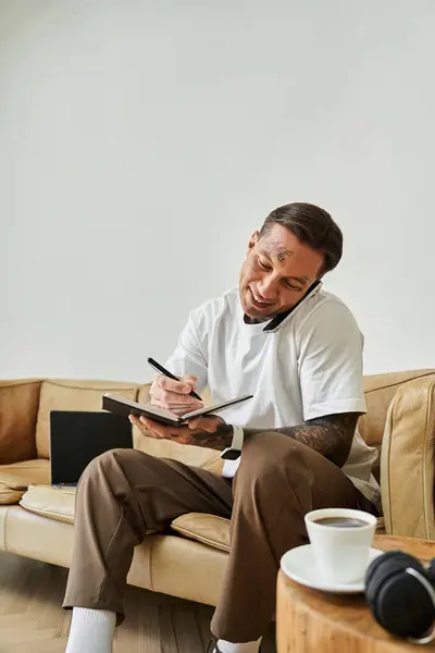 Un giovane uomo si siede comodamente a casa, facendo conversazioni telefoniche e prendendo appunti.. — Foto stock