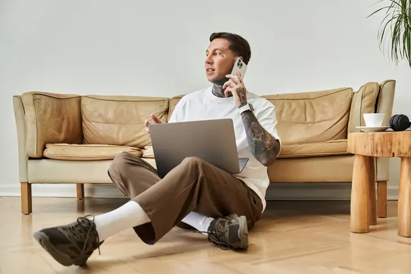 In a cozy living room, a young man sits on the floor with a laptop, talking on the phone. — Stock Photo