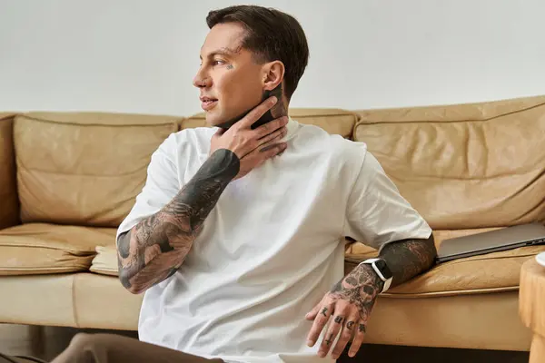 A young man sits comfortably in a stylish living room, thoughtfully touching his neck tattoo. — Stock Photo