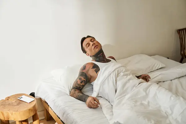 Un giovane uomo tatuato si sdraia a letto, assaporando una mattinata serena nel suo spazio accogliente. — Foto stock