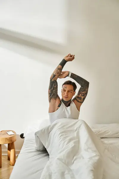 The young man with tattoos stretches his arms while waking up in a sunlit bedroom. — Stock Photo