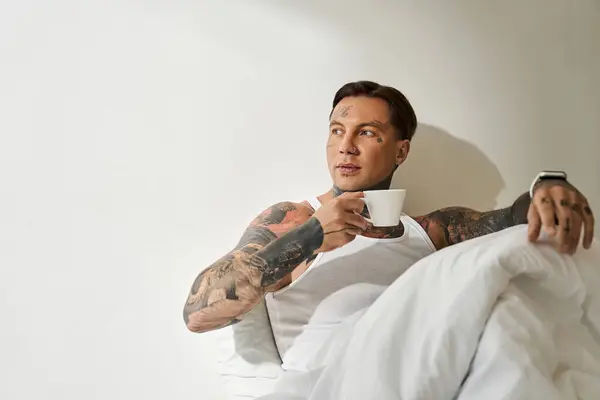 Un giovane uomo si rilassa comodamente a casa, sorseggiando caffè mentre è avvolto in una coperta. — Foto stock