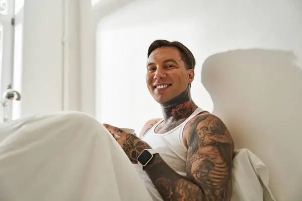 A young man sits comfortably in his bed, smiling brightly while enjoying his time indoors. — Stock Photo