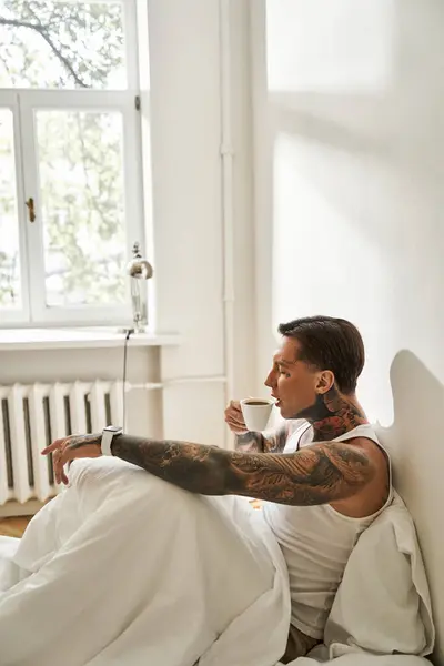 A young man with tattoos relaxes in bed, sipping coffee while soaking in the morning light. — Stock Photo