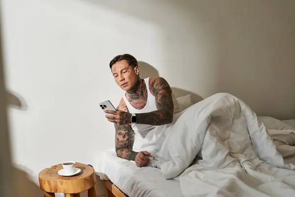 A handsome young man relaxes in bed, checking his phone with a cup of coffee nearby. — Stock Photo