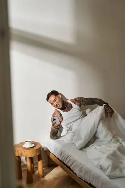 A handsome young man sits up in bed and smiles while checking his phone in morning sunlight. — Stock Photo