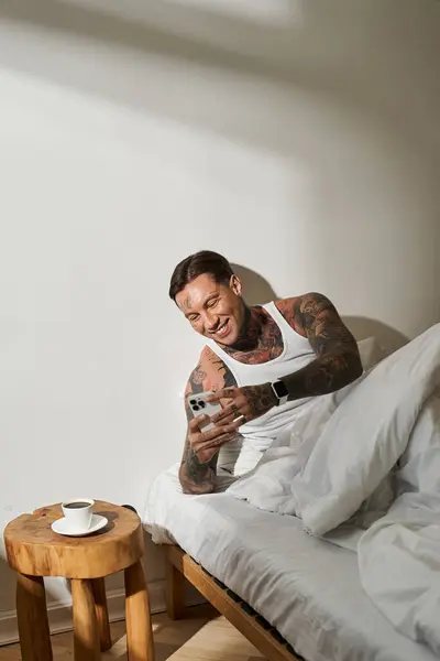 A young handsome man sits comfortably in bed, smiling at his phone while sipping coffee. — Stock Photo