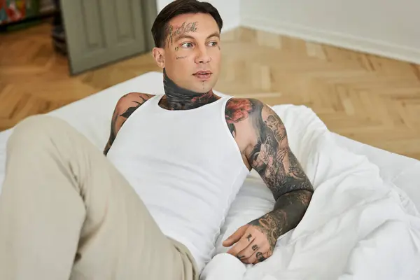A handsome young man lounges on a bed, displaying intricate tattoos and a casual style. — Stock Photo