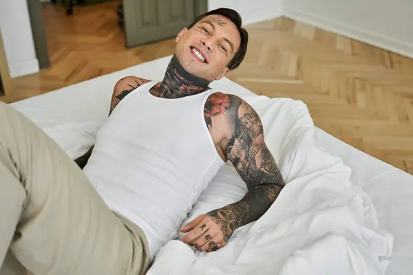 The young man lounges on a bed, radiating joy with his tattoos visible and a casual style. — Stock Photo