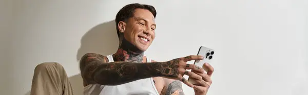A young, tattooed man enjoys a lighthearted moment while using his smartphone at home. — Stock Photo