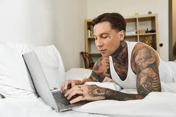 A young man with tattoos engages with his laptop while relaxing in bed at home. — Stock Photo