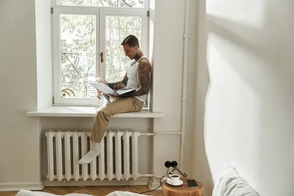 The young man sits on a windowsill, focused on his laptop as sunlight streams in. — Stock Photo