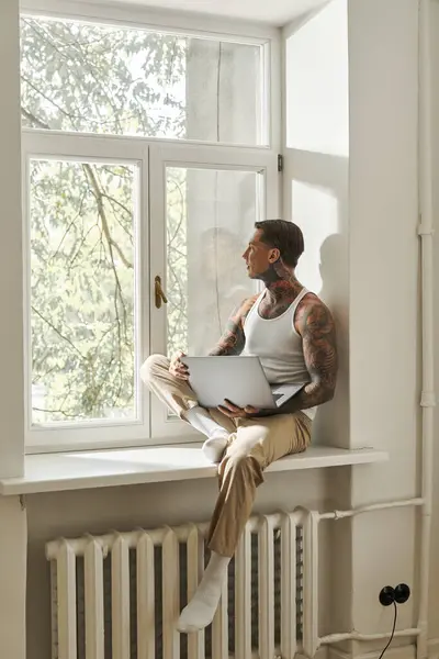 A young man sits comfortably on a windowsill, focused on his laptop as sunlight filters through. — Stock Photo