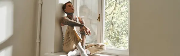 The young man, adorned with tattoos, enjoys a peaceful moment by the window, lost in thought. — Stock Photo