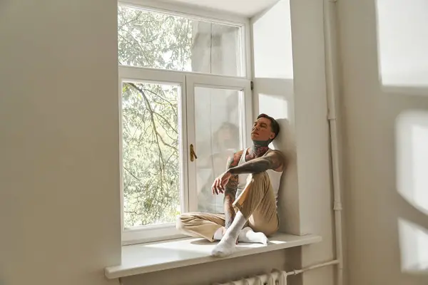 The young man sits comfortably on a windowsill, basking in warm sunlight with a relaxed demeanor. — Stock Photo