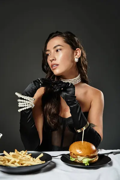 A chic woman in elegant attire enjoys her meal of a gourmet burger and fries while posing. — Stock Photo