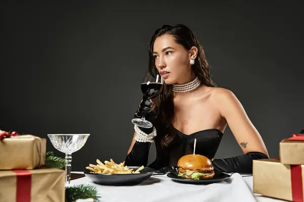 Uma jovem mulher em trajes chiques saboreia um hambúrguer gourmet e vinho em meio a presentes de férias. — Fotografia de Stock