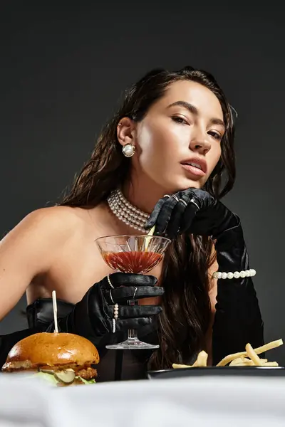 A sophisticated woman in elegant attire enjoys a cocktail alongside a gourmet burger and fries. — Stock Photo