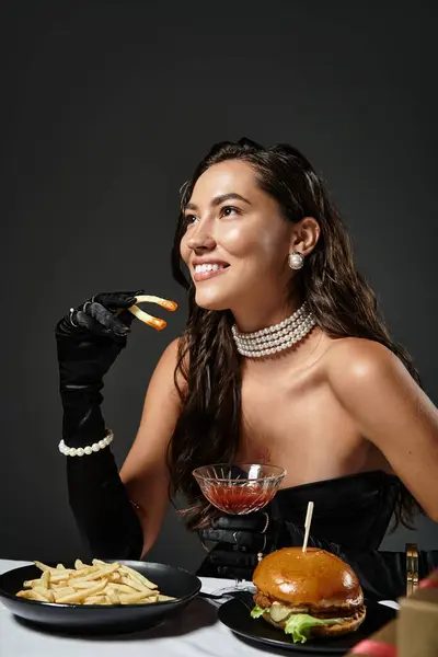 A sophisticated young woman in elegant attire savors gourmet fries and a burger while smiling. — Stock Photo