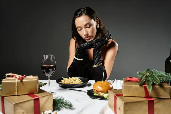 Una giovane donna in abiti eleganti assaporando un pasto gourmet circondato da regali di festa e vino. — Foto stock