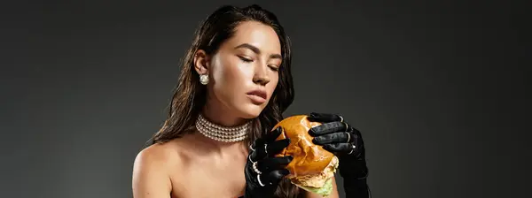 A stunning young woman in glamorous attire savors a gourmet burger with elegance and poise. — Stock Photo