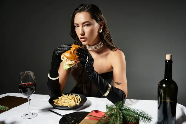 Una joven vestida con elegante atuendo saborea una hamburguesa gourmet mientras está sentada con buen vino y papas fritas. - foto de stock