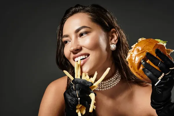 A stylish young woman playfully enjoying fries and a burger while dressed elegantly. — Stock Photo