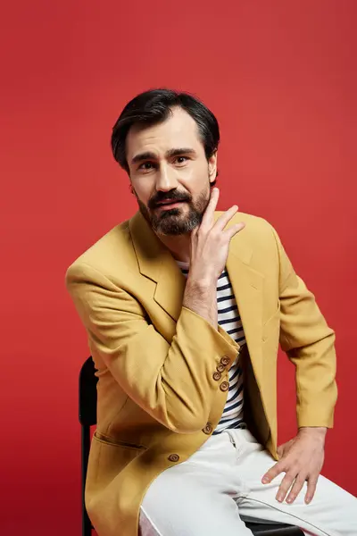 A middle aged man seated in a yellow blazer gently touches his chin, deep in thought. — Stock Photo