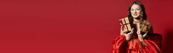 A cheerful young woman dressed in a stunning red gown presents two beautifully wrapped golden gifts. — Stock Photo