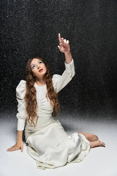 A young woman elegantly dressed in white gestures upwards, shimmering droplets surround her. — Stock Photo