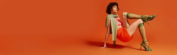 A young woman showcases her emotions while seated stylishly against a bold orange background. — Stock Photo
