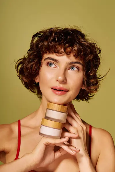 A young woman with curly hair displays two skincare jars while lost in thought and emotion. — Stock Photo