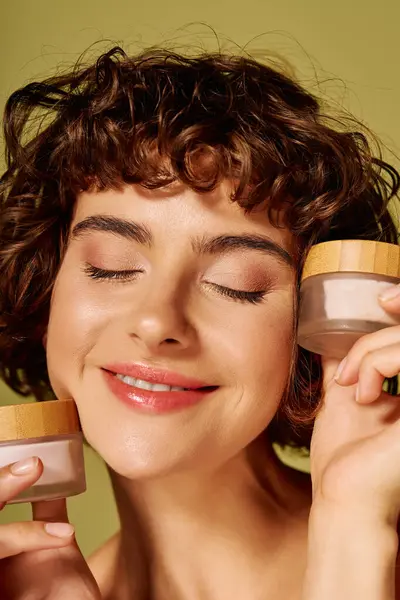 A young woman expresses happiness while holding skincare jars, radiating beauty and positivity. — Stock Photo