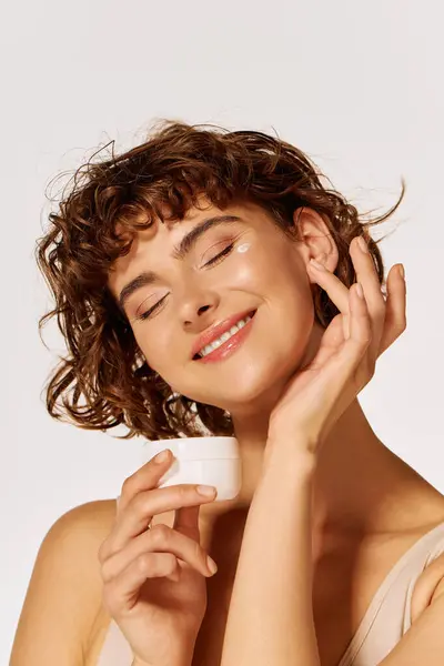 A joyful young woman enjoys a moment of happiness while holding a skincare jar gently. — Stock Photo