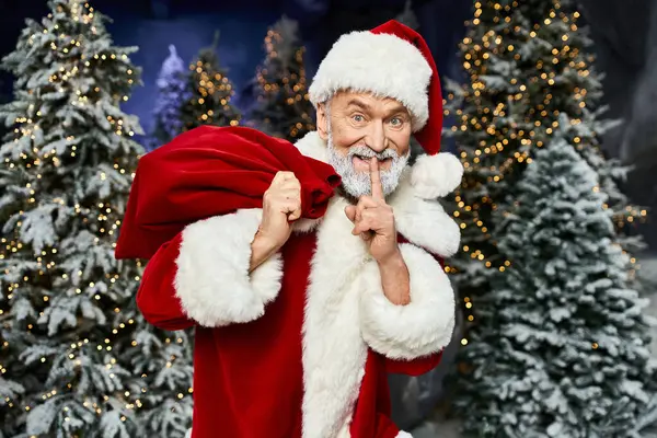 Papai Noel brinca gestos de silêncio enquanto segura um saco em meio a árvores cintilantes. — Fotografia de Stock