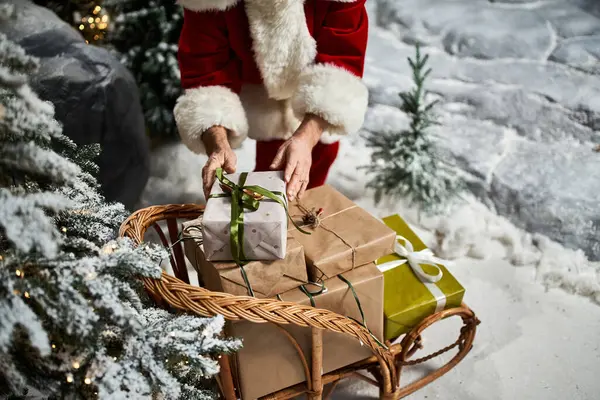 A cheerful figure in a red suit places wrapped gifts on a wooden sleigh surrounded by snow. — Stock Photo