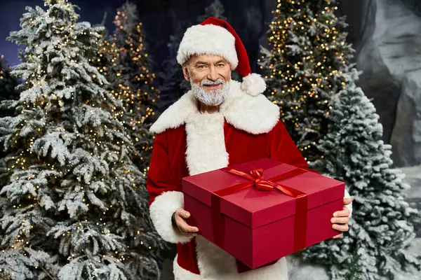 Santa warmly presents a beautifully wrapped gift amidst sparkling winter trees and twinkling lights. — Stock Photo