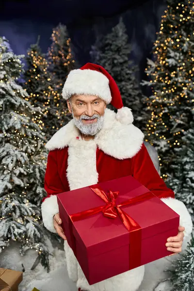 A festive Santa Claus holds a large red gift surrounded by snow covered trees and twinkling lights. — Stock Photo