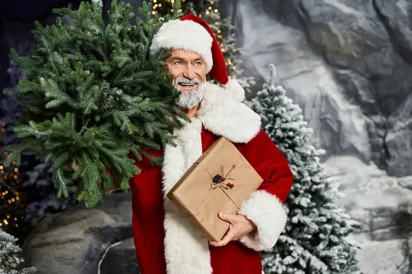 Santa Claus holds a festive gift while surrounded by snow covered trees and decorations — Stock Photo