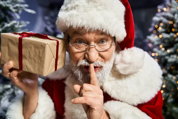 Santa Claus, with a wrapped gift, gestures playfully to keep a secret in the snow. — Stock Photo