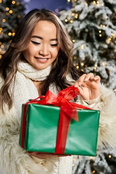 A young woman smiles as she unpacks a festive gift, surrounded by a snowy Christmas backdrop. — Stock Photo