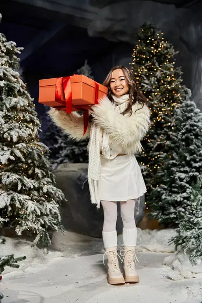 The woman beams while balancing a beautifully wrapped present amidst frosty trees and lights. — Stock Photo