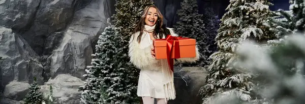 A joyful woman smiles while holding a gift box, surrounded by snowy trees and a winter wonderland. — Stock Photo