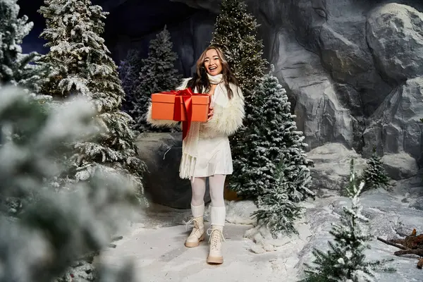A cheerful woman in a snowy landscape joyfully presents a vibrant red Christmas gift. — Stock Photo