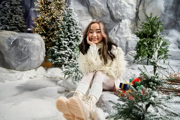Una mujer se sienta cómodamente en un paisaje nevado, rodeado de árboles de Navidad y decoraciones. - foto de stock