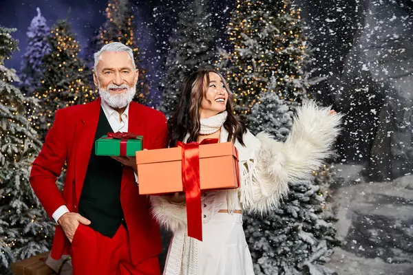 A cheerful couple embraces the Christmas spirit with colorful presents surrounded by lush trees. — Stock Photo